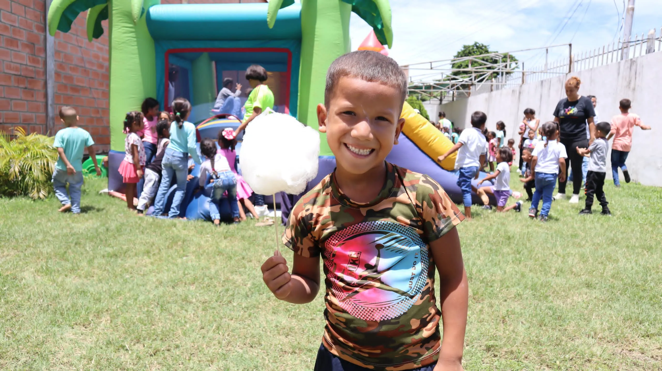 Smiling boy holding cotton candy with children playing near an inflatable bounce house outdoors.